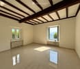 Empty room with exposed wooden beams on the ceiling, large windows offering a scenic view, tiled floor, and radiators under the windows.