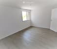 Empty room with light grey walls, wooden flooring, and a single window letting in natural light. The room has a white door on the right side.