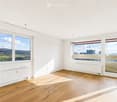 Empty room with wooden flooring, white walls, large windows offering views of both countryside and cityscape, radiators along the walls.
