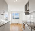 Modern kitchen with white cabinets, stainless steel appliances, wooden flooring, and large windows offering a city view.