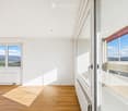 Bright, empty room with wooden flooring, white walls, large windows offering scenic views, and radiators along the walls.
