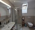 Modern bathroom with a white sink, mirror, shower stall, toilet, and a small wooden shelf holding toiletries. The room features a skylight window and a towel hanging on the wall.