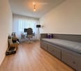 A modern bedroom with a bed, desk, chair, and lamp. The room features wooden flooring and large windows with white curtains.