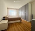 A modern bedroom with a large bed, a wardrobe, and a nightstand. The room features wooden flooring and large windows with curtains.