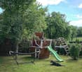 A well-maintained garden with a wooden playhouse, green slide, swing set, hammock, and a dog resting on the grass.