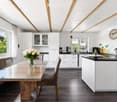 Modern kitchen with white cabinets, dark countertops, a wooden dining table with glass top, two chairs, and a bench. Features include a sink, faucet, coffee maker, blender, and a decorative vase with white roses on the table.