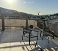 Balcony with tiled floor, metal railing, two chairs, small table, and scenic view of a town and hills.