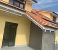 Exterior view of a modern two-story house with yellow walls, red-tiled roof, and a covered entrance area with a grey wooden garage.