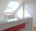 Bright, modern attic space with wooden flooring, white walls, and skylight windows allowing natural light. Features a white door and a sleek white counter with a red accent panel.