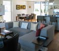 Modern living room with a grey sectional sofa, coffee table, dining area with chairs and table, kitchen island with bar stools, and decorative items including candles and artwork.