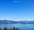 Panoramic view of a lake surrounded by mountains and a town, with clear blue skies and scattered trees in the foreground.