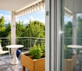 Balcony with a view of green trees, featuring a white chair, a white table, and a wooden planter with plants.