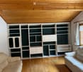 Living room with light wooden flooring, white built-in bookshelf with multiple compartments and cabinets, two beige sofas, and a window with white curtains.