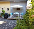A cozy outdoor patio area with a white table, four chairs, an umbrella, and a grill, surrounded by greenery and a stone-paved ground.