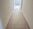 Clean, tiled hallway with light-colored walls and a smooth floor, leading to an open area with large windows and a view of a balcony.