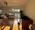 Bright living room with wooden flooring, large sliding glass doors leading to a balcony, a sofa, a small table, a chair, a floor lamp, and a potted plant.