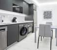 Modern kitchen and dining area with stainless steel appliances, white countertops, and grey cabinets. Includes a sink, coffee maker, dishwasher, washing machine, oven, refrigerator, dining table, and chairs.