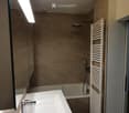 Modern bathroom with a white sink, chrome faucet, bathtub, showerhead, and a white radiator. The walls are tiled in beige, and the ceiling has a round light fixture.