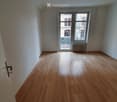 Empty room with wooden flooring, white walls, and a large window with a view of a building and balcony.
