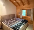 Cozy attic bedroom with wooden beams and slanted ceilings, featuring a large double bed with patterned bedding, a nightstand with a lamp, and large windows with curtains.