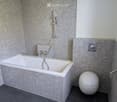 Modern bathroom with a white bathtub, toilet, and showerhead, featuring grey tiled walls and dark tiled floor.