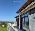 Modern two-story building with white siding, large glass windows, and a wooden roof. Features a balcony with a metal railing and a scenic view of the surrounding landscape.