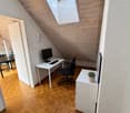 A modern, well-lit attic room with wooden herringbone flooring, white walls, and a slanted wooden ceiling. Features include a desk with a computer, a comfortable office chair, a small side table with a potted plant, and a large skylight providing natural