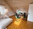 Modern attic room with wooden slanted ceilings, herringbone wooden flooring, a white sofa, a round dining table with green chairs, a floor lamp, and a potted plant on the table.