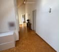 A narrow hallway with wooden herringbone flooring, white walls, and several doors leading to other rooms. A small table with a vase of flowers is visible at the end of the hallway.