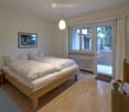 Cozy bedroom with a wooden bed, nightstand, and large windows leading to a balcony. The room features light wooden flooring and a picture frame on the wall.