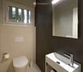 Modern bathroom with white toilet, white sink with chrome faucet, large mirror, and built-in storage. Light-colored walls and floor, with a small window providing natural light.