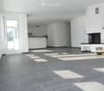 Empty room with grey tiled floor, white walls, and large windows allowing natural light. Features a fireplace and a kitchen area with cabinets and a countertop.