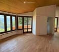 Empty room with wooden flooring, large windows, and a view of a forested area. The room has a slanted wooden ceiling and a bench near the windows.