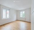 Completely empty room with bare white walls, wooden floor, and two windows with white frames. No furniture or equipment.
