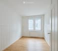 Empty room with light wooden flooring, white walls, and a large window with white frames. The walls feature a wainscoting design below the main wall surface.