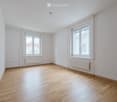 Empty room with light wooden flooring, white walls, and two windows with radiators beneath them.
