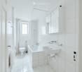 Modern, white-tiled bathroom with a toilet, bathtub, sink, mirror, and cabinet.