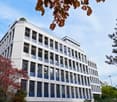 modern white and blue building, 5 floors, numerous windows, glass balcony on top floor, surrounded by trees