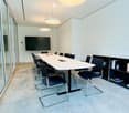 Modern conference room with a long white table, black chairs, a large screen on the wall, and a bookshelf with books and magazines.