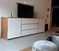 Modern living room with a grey ottoman, a sleek white and wood TV stand, a flat-screen TV, and decorative vases on the stand.