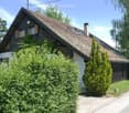 Traditional wooden house with a steeply pitched roof, surrounded by greenery and bushes.