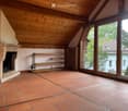 An unfurnished room with exposed wooden beams, terracotta tiles, a fireplace, and large windows offering a view of the outdoors.