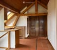 A spacious, well-lit hallway with exposed wooden beams and a high, slanted ceiling. The floor is covered in terracotta tiles, and the walls are painted white. There are two wooden double doors at the end of the hallway, and a wooden bench or table is posi