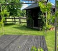 A serene garden area with a wooden deck, lush green grass, and a small gazebo surrounded by climbing plants and potted flowers.