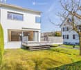 Modern white house with glass windows and a wooden deck, surrounded by a well-maintained garden.