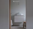 Modern bathroom with a white sink, cabinet, and wooden stool. Light grey tiled walls and a concrete floor.
