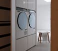A modern, minimalist laundry area with a built-in washing machine and dryer unit, a small wooden stool with a folded towel, and a bright, airy space with natural light from a skylight.
