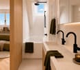 Modern, minimalist bathroom with a green sofa, wooden floor, large windows, a bathtub, and a double sink with modern faucets.