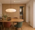 Modern dining area with a round wooden table, four chairs, a vase with flowers, two bottles, and a bowl on the table. A pendant light hangs above, and a doorway leads to a bathroom.