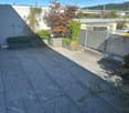 Roof terrace with grey tiled flooring, potted plants, and a metal fence, overlooking a cityscape with buildings and trees.